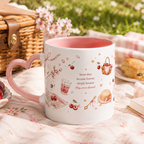 A white ceramic mug with a pink heart-shaped handle and pink interior, decorated with cherry and picnic illustrations and the phrase 'Some days become favorite simply because they were shared.' It is shown on a pink picnic blanket outdoors with a wicker basket, cherries, and scones in the background.