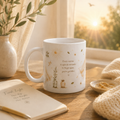 A white ceramic mug with delicate botanical illustrations and the quote 'Every sunrise is a gentle reminder to begin again you've got this' sits on a wooden table in a warm, sunlit room setting next to an open notebook, a knit blanket, and a vase.