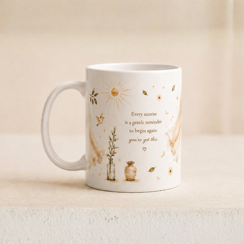 A white ceramic mug with a handle, decorated with gold-toned illustrations of a sun, leaves, birds, and a small vase, alongside the printed quote: 'Every sunrise is a gentle reminder to begin again you've got this.' The mug sits on a light beige surface against a softly blurred neutral background.