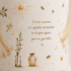 A close-up of a cream-colored cylindrical container or candle with a warm-toned printed design. The design features the quote 'Every sunrise is a gentle reminder to begin again.