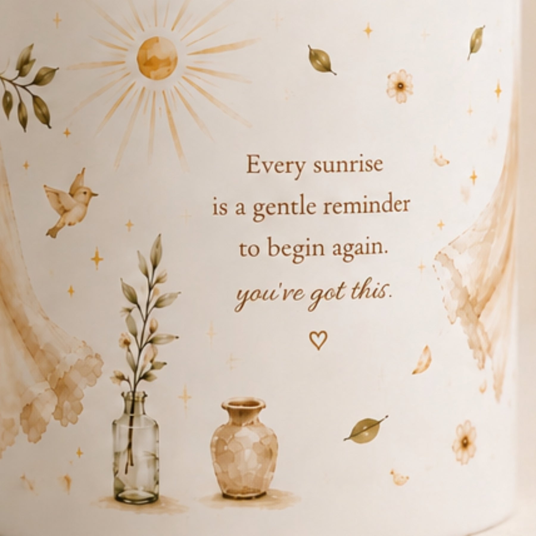 A close-up of a cream-colored cylindrical container or candle with a warm-toned printed design. The design features the quote 'Every sunrise is a gentle reminder to begin again.