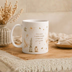 A white ceramic mug with a delicate gold and brown botanical illustration and the quote 'Every sunrise is a gentle reminder to begin again you're got that' sitting on a wooden surface in a cozy, warm-toned setting.