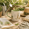 A ceramic mug with a green handle and interior, decorated with illustrations of potted plants and the quote 'Find joy in the little things that make you feel content.' It is styled on a wooden table with gardening props, including a watering can, gloves, a hat, and an open book, against a soft-focus garden background.