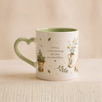 A white ceramic mug with a light green heart-shaped handle and interior, decorated with delicate botanical illustrations and the quote 'Find joy in the little things that make your heart bloom.'.