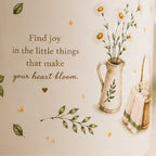 A cream-colored wall decal or art print featuring a cozy, vintage-style illustration. The design includes the quote 'Find joy in the little things that make your heart bloom.