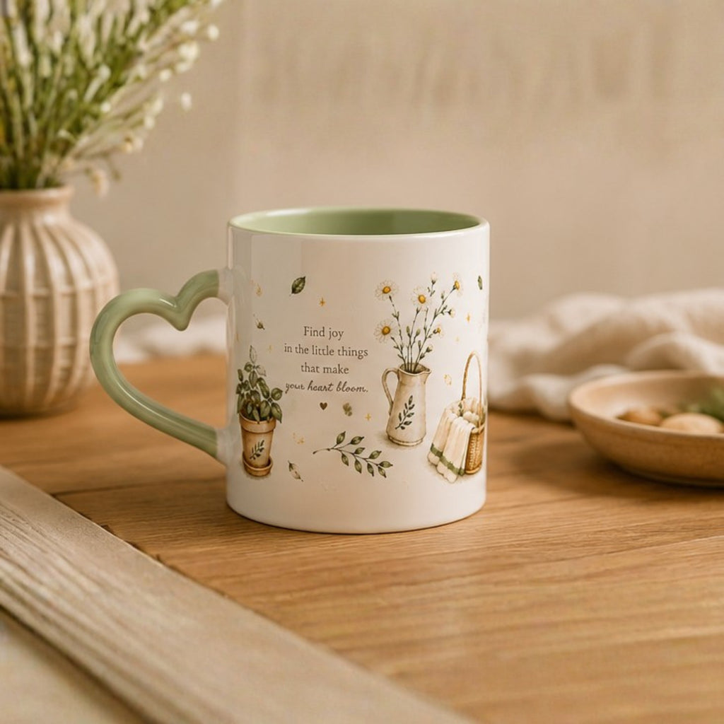 A ceramic mug with a heart-shaped handle sits on a wooden table. The mug is white with a light green interior and handle, decorated with illustrations of small potted plants, a watering can, and the text 'Find joy in the little things that make you happy most'.