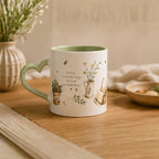 A ceramic mug with a heart-shaped handle sits on a wooden table. The mug is white with a light green interior and handle, decorated with illustrations of small potted plants, a watering can, and the text 'Find joy in the little things that make you happy most'.