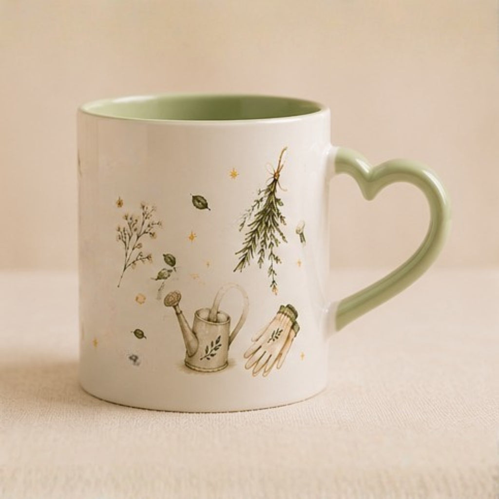 A white ceramic mug with a heart-shaped handle, featuring delicate botanical illustrations of a watering can, a hand, and a pine tree, with a light green interior and handle.