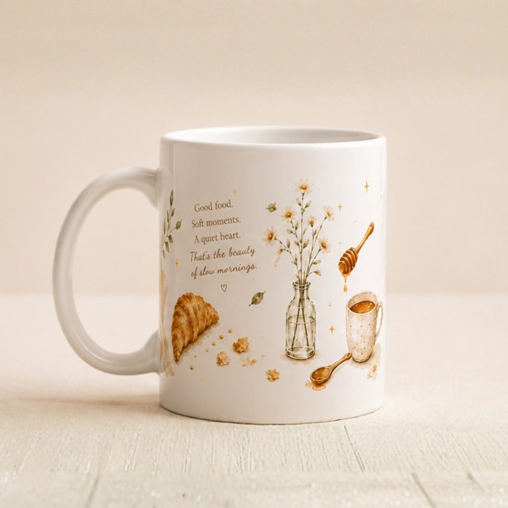 A white ceramic coffee mug with a warm brown and gold illustration of a croissant, honey, a small vase with wildflowers, and a teacup, alongside the quote 'Good food, soft moments. A quiet heart.