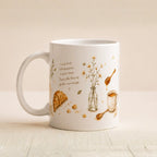 A white ceramic coffee mug with a warm brown and gold illustration of a croissant, honey, a small vase with wildflowers, and a teacup, alongside the quote 'Good food, soft moments. A quiet heart.