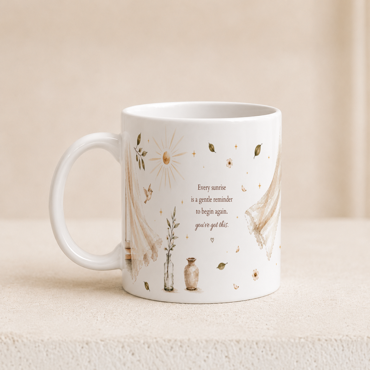 A white ceramic mug with a delicate beige and brown illustration of a sun, flowers, leaves, and lace, featuring the quote 'Every sunrise is a gentle reminder to begin again, you've got this.' It is set on a textured beige surface against a soft beige background.